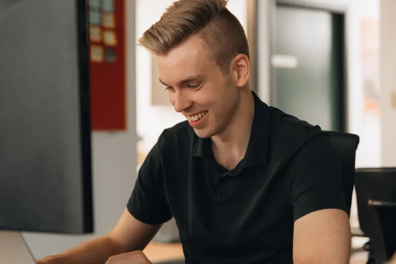 A young man in a black polo smiling at his desk as he works