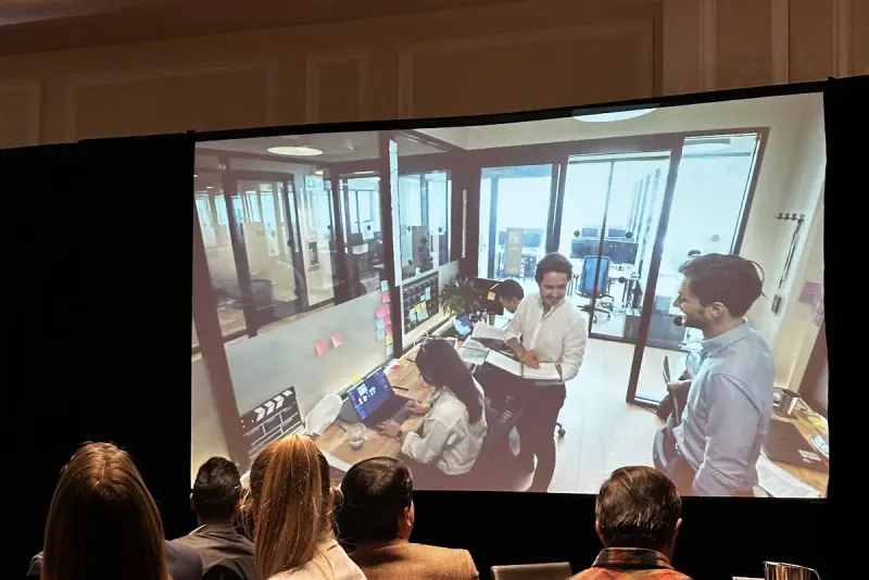 An audience watching a projected video of a team meeting in a glass-walled office