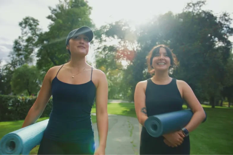 Two women walking with yoga mats through a sunlit park