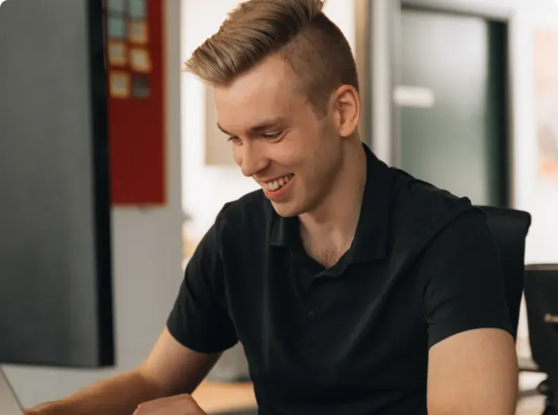 A young man in a black polo smiling at his desk